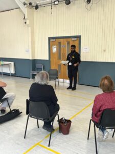 a presenter speaks to a group of elderly people sitting on chairs in a hall